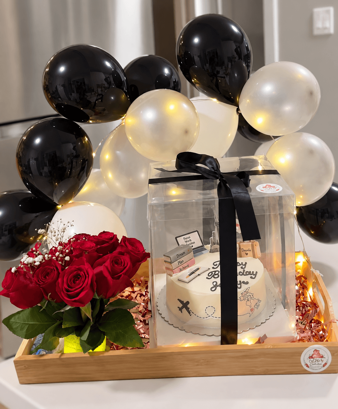 Nova Cakes gift hamper on a decorative wooden tray with black and cream balloons, fairy lights, a fresh red rose bouquet and a 100% eggless birthday cake in a clear ribbon-tied box, Halifax
